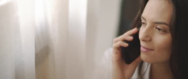 Beyaz gömlekli bir kadının telefonda biriyle konuşurken, gülümserken, pencerenin yanında bir yatakta otururken. Ağır çekim, BMPCC 4K