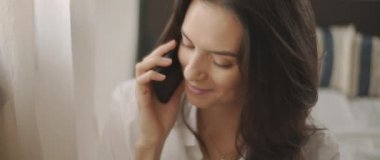 Beyaz gömlekli bir kadının telefonda biriyle konuşurken, gülümserken, pencerenin yanında bir yatakta otururken. Ağır çekim, BMPCC 4K