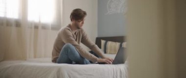 Genç bir adam yatakta oturuyor, dizüstü bilgisayarda yazıyor, evden çalışıyor. Yavaş çekim, orta çekim, BMPCC 4K