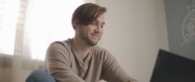 Yatakta oturup bilgisayar başında çalışırken gülümseyen genç bir adamın yakın çekimi. Ağır çekim, BMPCC 4K