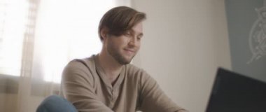 Yatakta oturup bilgisayar başında çalışırken gülümseyen genç bir adamın yakın çekimi. Ağır çekim, BMPCC 4K