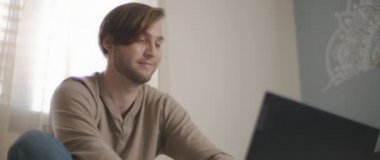 Yatakta oturup bilgisayar başında çalışırken gülümseyen genç bir adamın yakın çekimi. Ağır çekim, BMPCC 4K