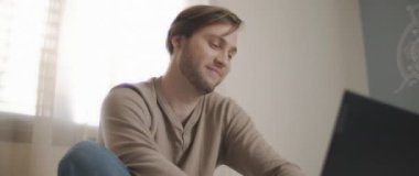 Yatakta oturup bilgisayar başında çalışırken gülümseyen genç bir adamın yakın çekimi. Ağır çekim, BMPCC 4K