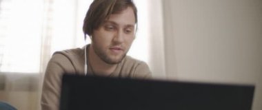 Laptopuyla uğraşırken kulaklıkla konuşan genç bir adam. Ev kavramından çalış. Ağır çekim, BMPCC 4K