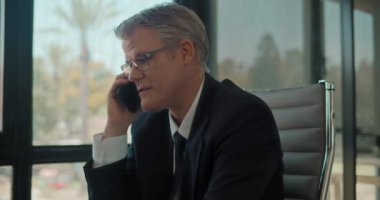 Resmi giyinen ve gözlüklü bir işadamı ofiste çalışırken telefonla konuşuyor. İzleme çekimi, yavaş çekim.. 