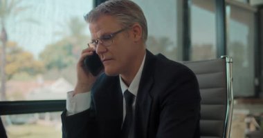 Resmi takım elbiseli ve gözlüklü bir işadamı ofiste çalışırken telefonla konuşuyor, gülümsüyor. İzleme çekimi, yavaş çekim.. 
