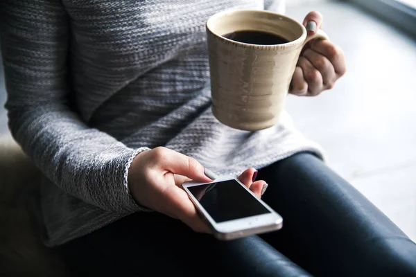 Belle fille élégante en pantalon en cuir et un pull moderne assis sur le sol avec une tasse de café et téléphone mobile — Photo