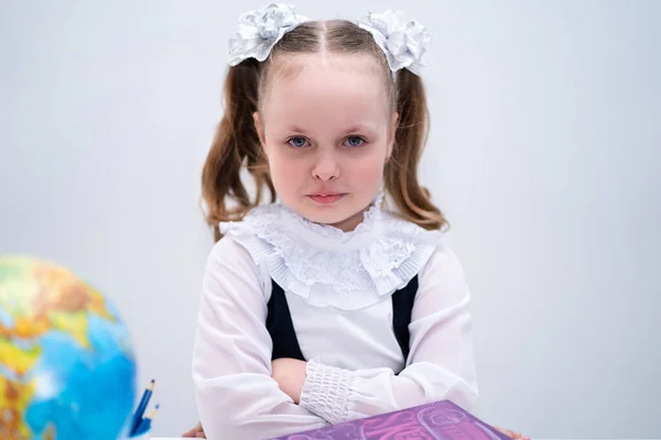 A girl is sitting at a table on a white background, in a school uniform, with white bows and wants to cry