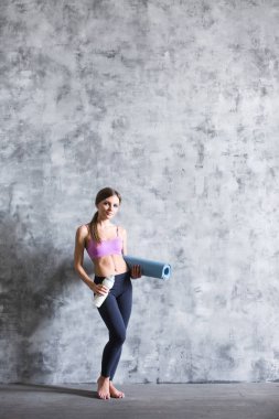 Spor giyim güzel genç kadın bir yoga mat ve bir şişe su tutarak, kameraya bakıyor ve gülümsüyor, gri bir arka plan üzerinde duran.