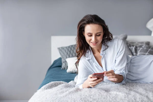 Mulher encantadora usando celular enquanto deitada na cama aconchegante em casa pela manhã . — Fotografia de Stock