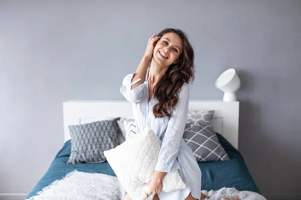 Mujer positiva con almohada sentada en la cama y sonriendo a la cámara en casa. — Foto de Stock