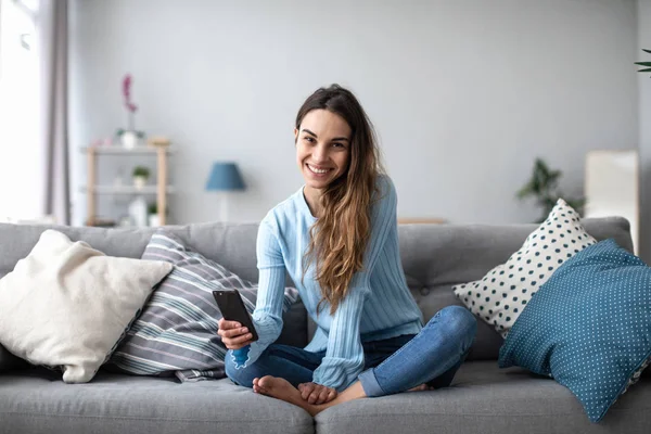 Attraktiv leende kvinna med hjälp av smart telefon när du sitter på soffan hemma. — Stockfoto
