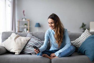 Modern akıllı telefon kullanarak evde gülümseyen genç bir kadın. Çevrim içi sohbet.