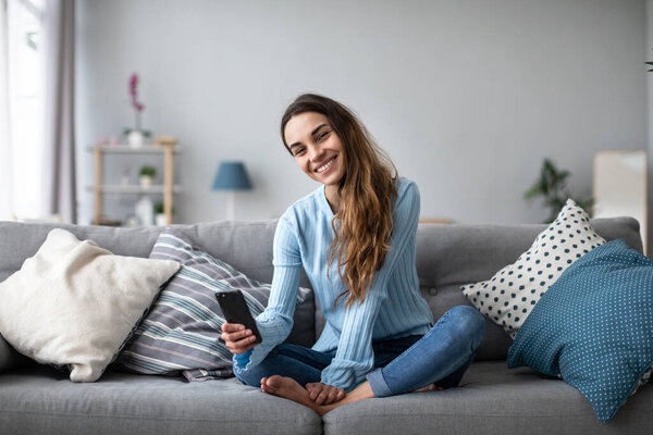 Online chat. Smiling woman talking on smartphone with friends while sitting on sofa at home. Portrait of a positive girl indoors.