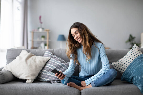 Онлайн чат. Улыбающаяся женщина разговаривает по смартфону с друзьями, сидя дома на диване. Портрет позитивной девушки в помещении
.