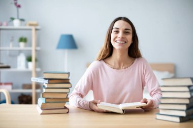 Neşeli genç bayan kitaplarla dolu bir masada oturuyor ve ödev yapıyor. Evde öğreniyorum..