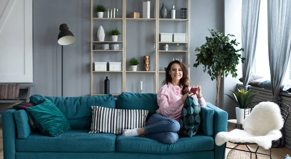 Oturma odasındaki koltukta akıllı telefonuyla gülümseyen güzel yalnız kadın. Evde cep telefonunda kullan.