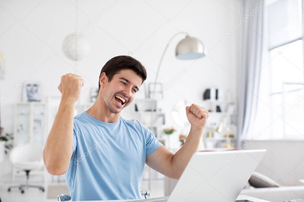 Joyful young man in a blue t-shirt with a laptop rejoices looking at the screen. Cheerful freelance, distance job.