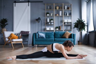 Spor kıyafetli genç bir kadın evde yoga minderinde yerde otururken esneme hareketleri yapıyor. Spor ve spor.