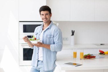 Salata tabağı dolusu taze sebzeli yakışıklı gülümseyen genç adam kameraya bakıyor. Mutfakta mutlu sıradan bir adamın portresi..