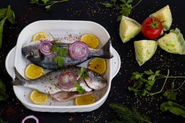 Siyah yüzey üzerinde dilimlenmiş soğan ve limonla birlikte iki adet terbiye edilmiş çipura pişirmeye hazır..