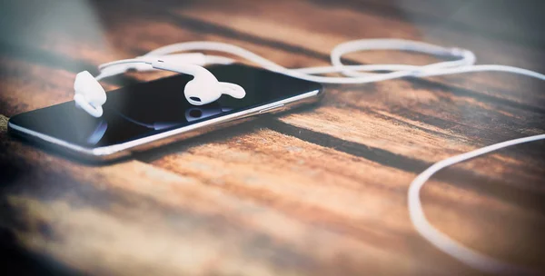 A 45 degree view of a set of wired earbud headphones and mobile phone lying on a rustic wooden table. Styling and grain effect added to image.