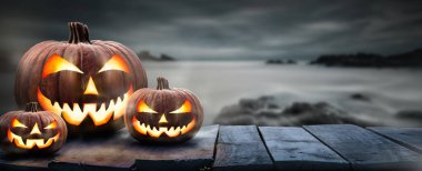 Üç ürkütücü cadılar bayramı balkabağı, kötü yüzlü ve tahta bir bankta gözleri olan Jack O Lantern. Ürün yerleştirmek için yeri olan sisli, gri bir sahil gece arka planına sahip bir masa..