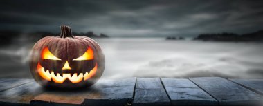 Korkunç bir cadılar bayramı balkabağı, Jack O Lantern, tahtadan bir bankta şeytani bir yüzü ve gözleri olan, sisli gri bir kıyı gecesi geçmişi olan, ürün yerleştirme alanı olan bir masa.