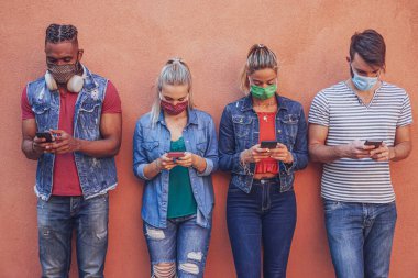  Yüz maskesi takarak akıllı telefonlarını 19 kez kullanan bir grup insan, duvarın yanında durup cep telefonlarını tutarken internet haberlerini kontrol eden arkadaşlar.
