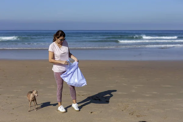 Frau mit Hund sammelt Müll und Plastik am Strand auf — Stockfoto