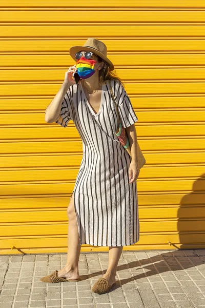 Middle-aged woman wearing a hat, sunglasses and a rainbow-colored protective mask. LGTB on yellow background. Covid-19 concept