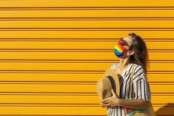 Middle-aged woman wearing a hat, sunglasses and a rainbow-colored protective mask. LGTB on yellow background. Covid-19 concept