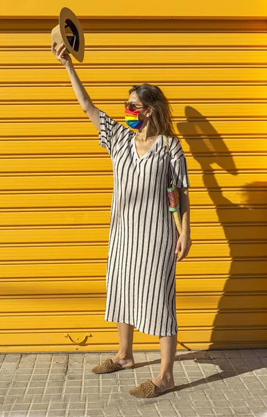 Middle-aged woman wearing a hat, sunglasses and a rainbow-colored protective mask. LGTB on yellow background. Covid-19 concept