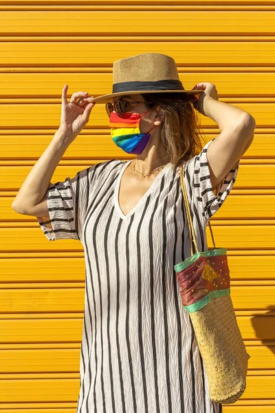 Middle-aged woman wearing a hat, sunglasses and a rainbow-colored protective mask. LGTB on yellow background. Covid-19 concept