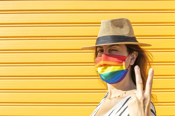 Middle-aged woman wearing a hat, sunglasses and a rainbow-colored protective mask. LGTB on yellow background. Covid-19 concept