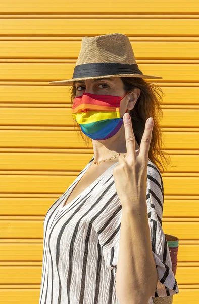 Middle-aged woman wearing a hat, sunglasses and a rainbow-colored protective mask. LGTB on yellow background. Covid-19 concept