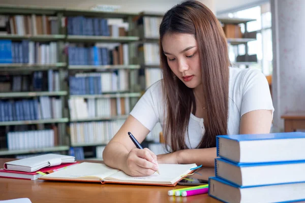 Asyalı bir kız öğrenci masada oturuyor ve bir deftere yazı yazıyor. Genç kız öğrenciler kütüphanede ödev yapıyor.