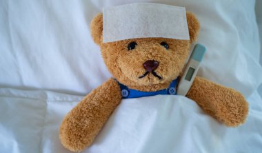 Brown teddy bear with fever reducing sheet and body temperature monitor. Used for health insurance or illness concepts clipart