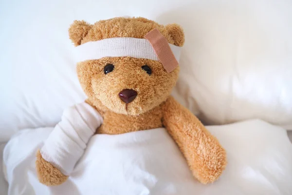Teddy bear is sick on the bed, his arm is broken and his head is broken from an accident.