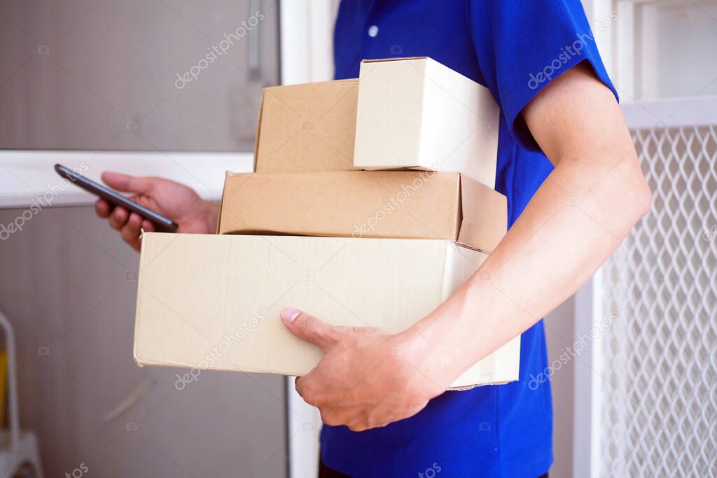 The shipper in the blue suit holds a brown box and smartphone standing in front of the customer's house. Free shipping for online shopping and express delivery