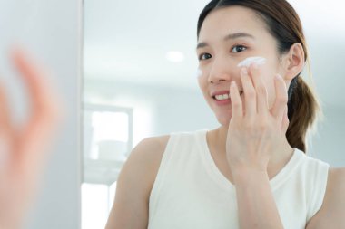 Güzel bir Asyalı kadın aynanın önünde oturur. Krem sürerek makyaj yapan sağlıklı bir kadın yüzü. Cilt kremi reklamı, kırışıklık önleyici, bebek yüzü, beyazlatma, nemlendirici, gözenekleri sıkılaştırma serumu.