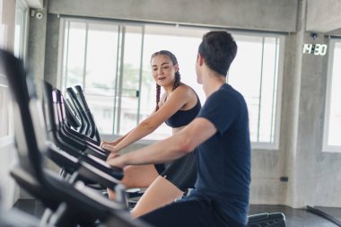 Erkek ve bayan sporcular spor salonunda egzersiz yapmaktan, bisiklete binmekten bahsederler. Spor kıyafetleri giyerler..