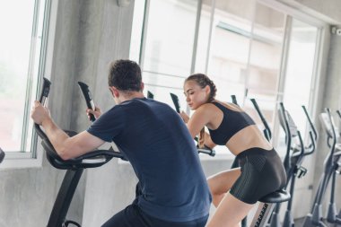 Erkek ve bayan sporcular spor salonunda egzersiz yapmaktan, bisiklete binmekten bahsederler. Spor kıyafetleri giyerler..