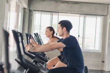 Erkek ve bayan sporcular spor salonunda egzersiz yapmaktan, bisiklete binmekten bahsederler. Spor kıyafetleri giyerler..