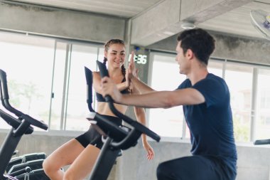 Erkek ve bayan sporcular spor salonunda egzersiz yapmaktan, bisiklete binmekten bahsederler. Spor kıyafetleri giyerler..