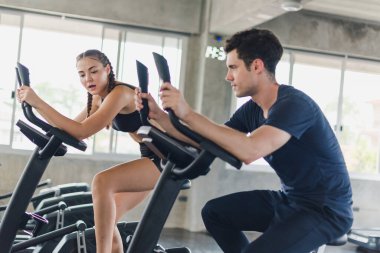 Erkek ve bayan sporcular spor salonunda egzersiz yapmaktan, bisiklete binmekten bahsederler. Spor kıyafetleri giyerler..