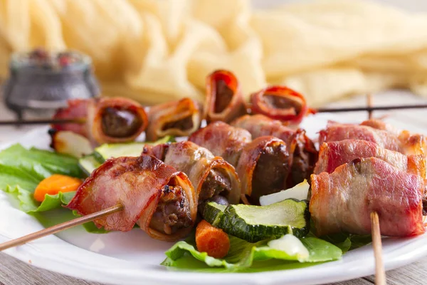 Chicken liver wrapped with bacon on skewers. Grilled liver kebabs with vegetables