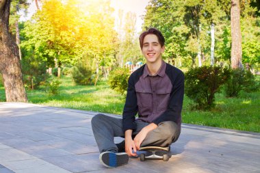 Oturma ve Şehir Parkı içinde buz pateni sonra kaykay üzerinde rahatlatıcı genç adam. Genç çocuk patenci