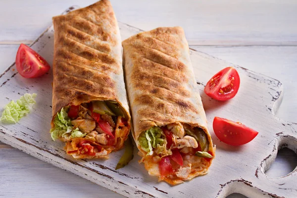 Chicken wraps with tomatoes, pickles, cabbage and onion on white chopping board and wooden table. Tortilla, burritos, sandwiches, twisted rolls