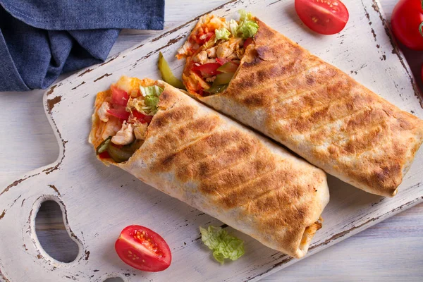 Chicken wraps with tomatoes, pickles, cabbage and onion on white chopping board and wooden table. Tortilla, burritos, sandwiches, twisted rolls